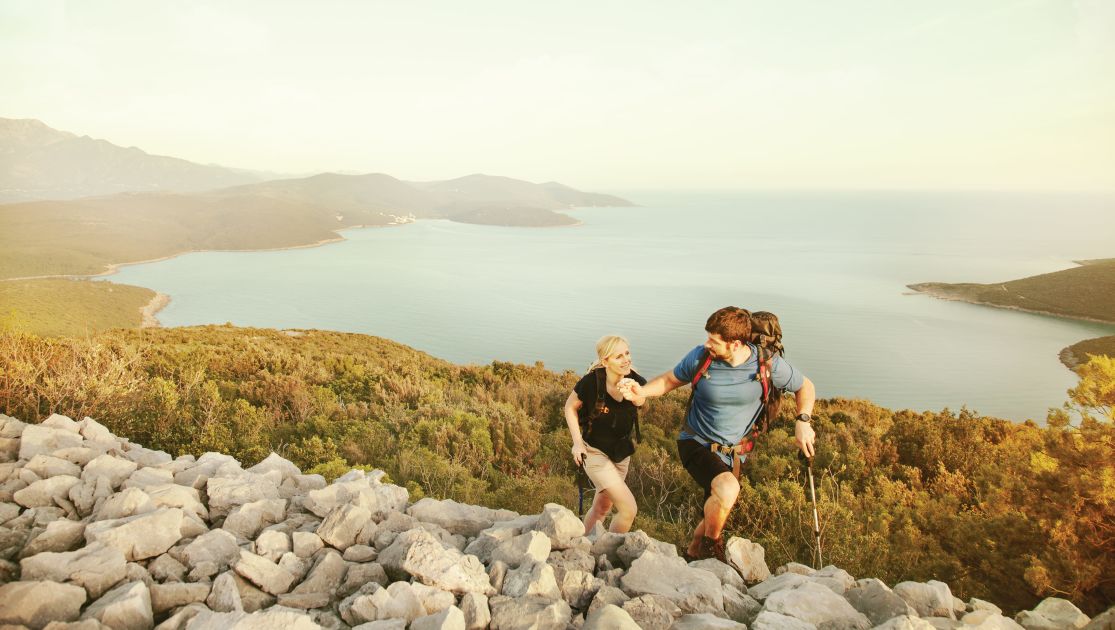 A Couple Of People Hiking On A Rocky Hill By A Body Of Water