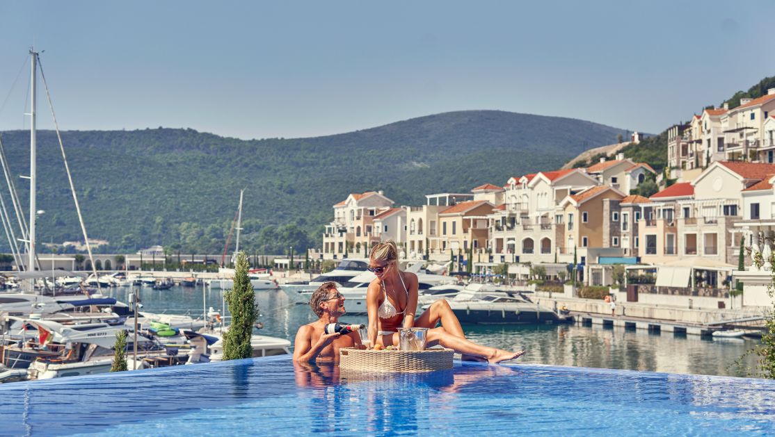 A Couple Of Women Sitting In A Pool By A Dock With Boats And Buildings And Mountains In The