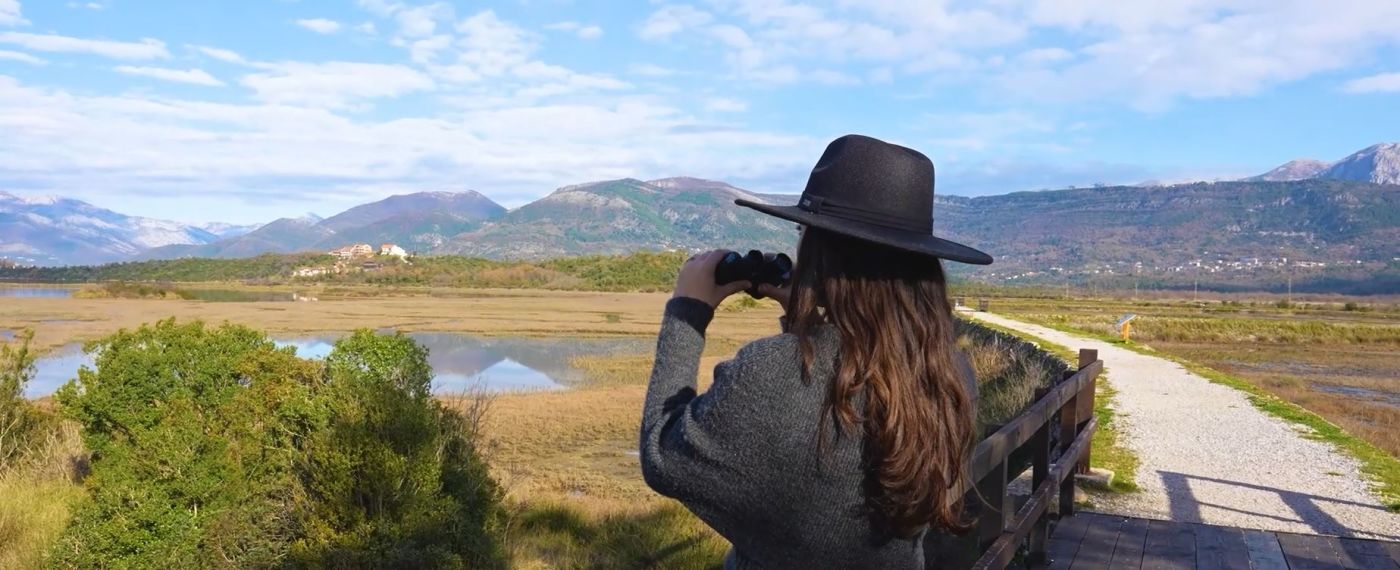 A Woman Birdwatching Over A River & Mountains Landscape